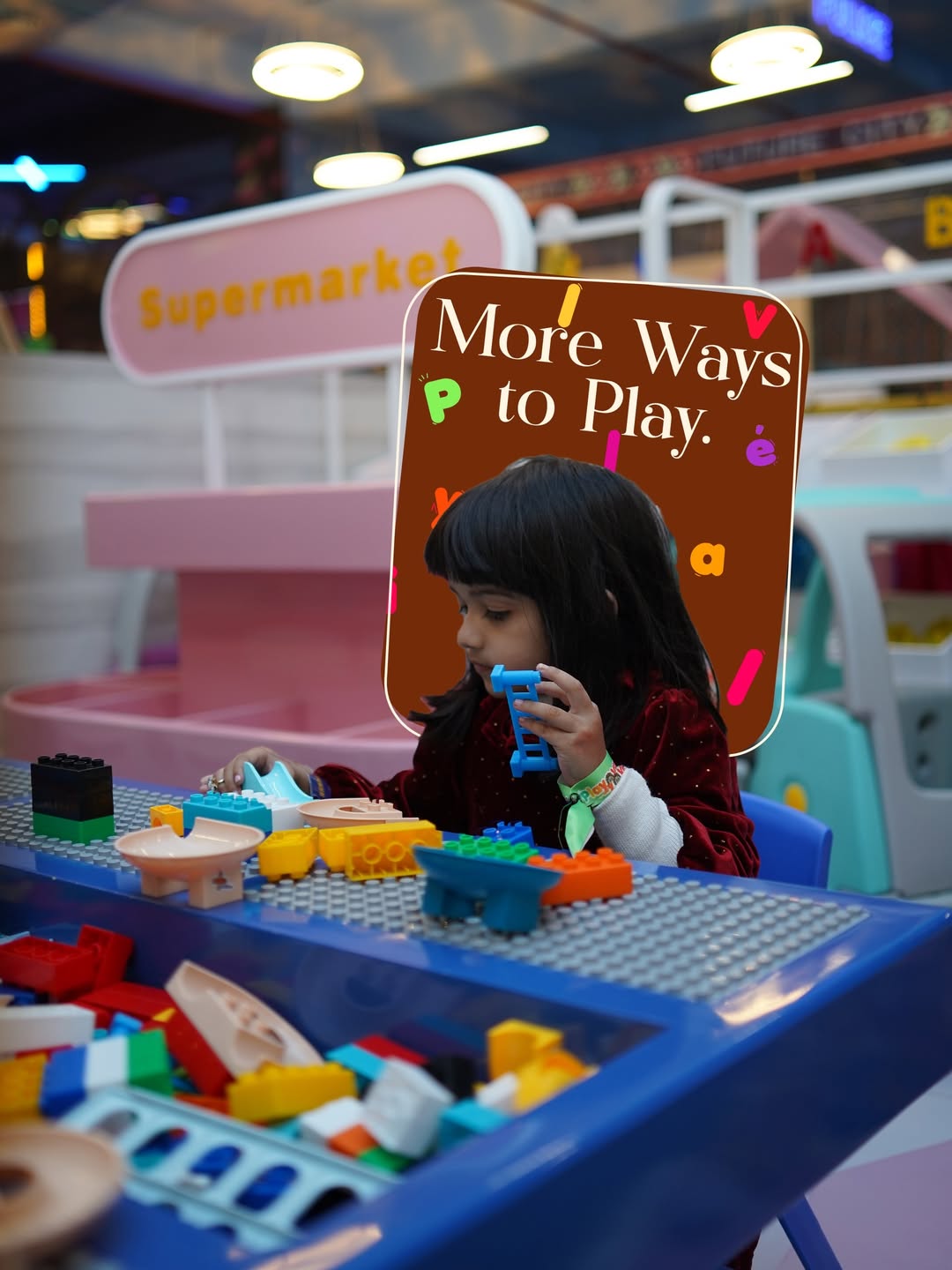 A young girl plays with colorful building blocks at a toy table in a brightly lit indoor play area.
