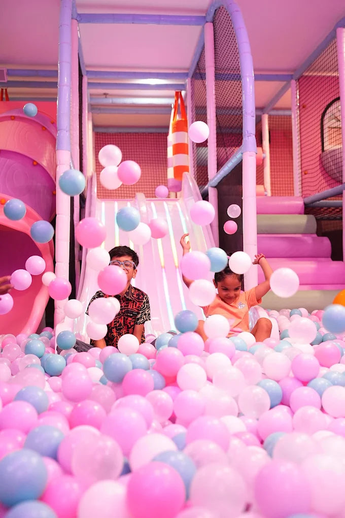 Children play in a ball pit filled with pink and blue balls, with a soft play structure in the background.