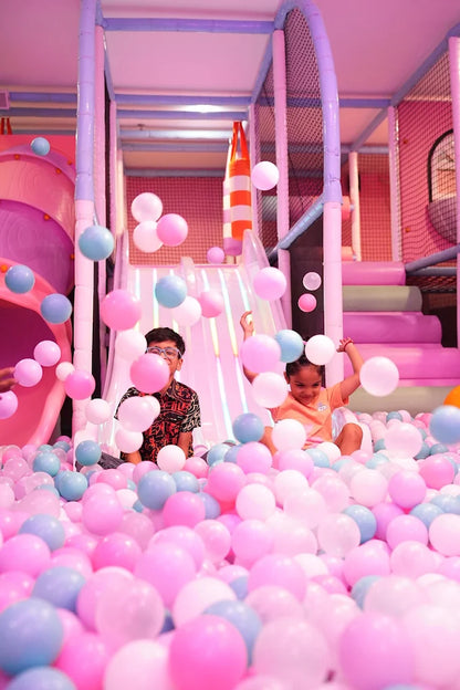 Children play in a ball pit filled with pink and blue balls, with a soft play structure in the background.