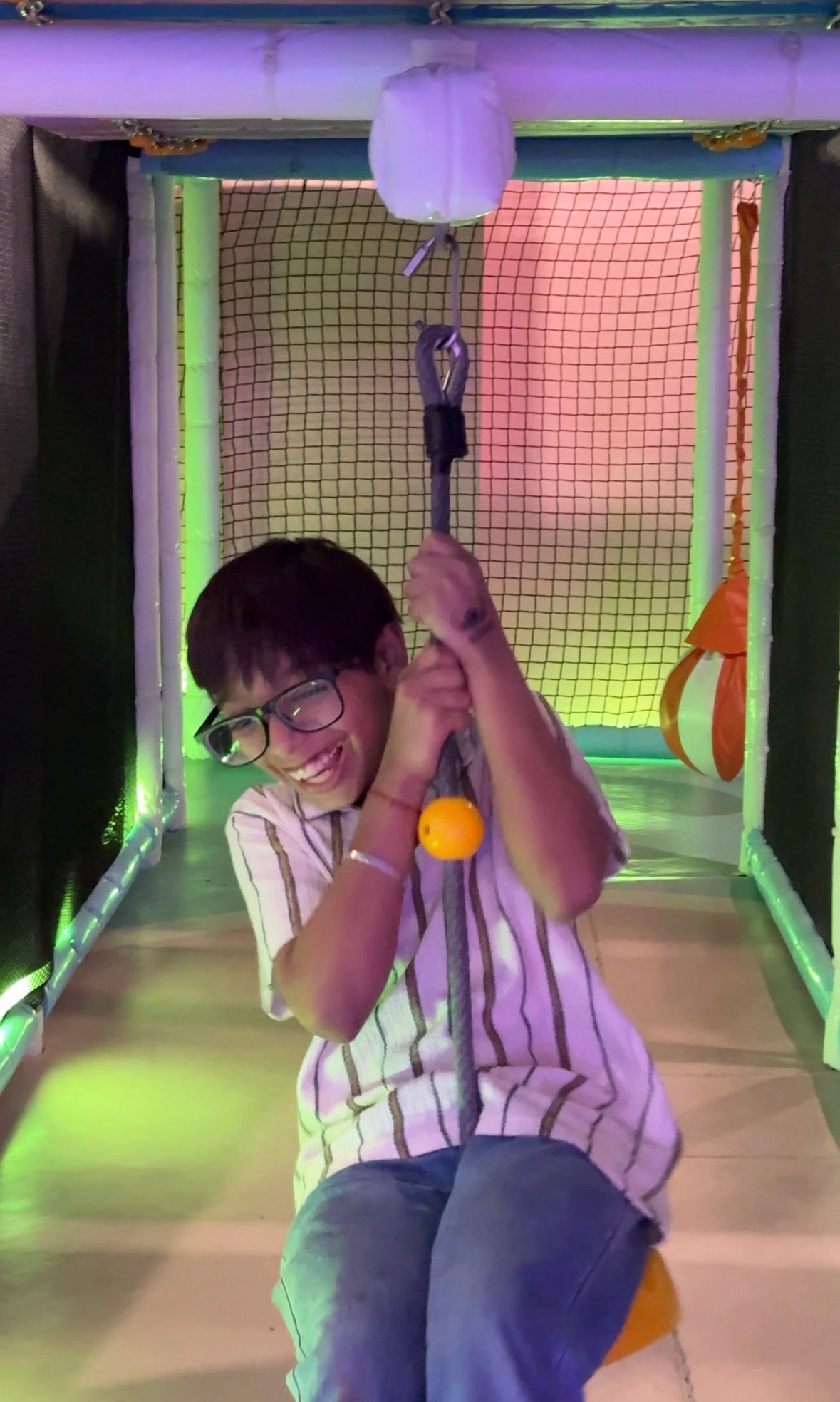 A young person laughs while ziplining through a colorful indoor play area.