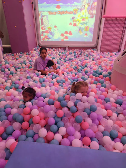 Children play in a ball pit with a festive screen in the background.