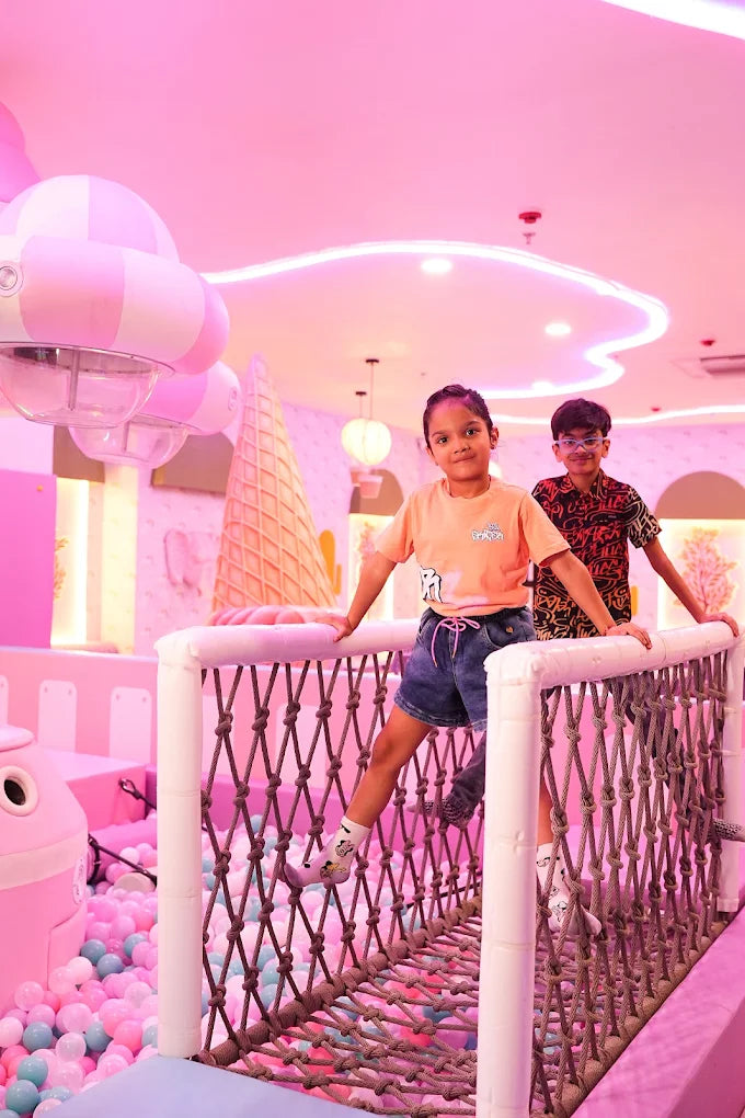 Two children stand on a rope bridge in a pink, ice cream-themed play area filled with ball pit balls.