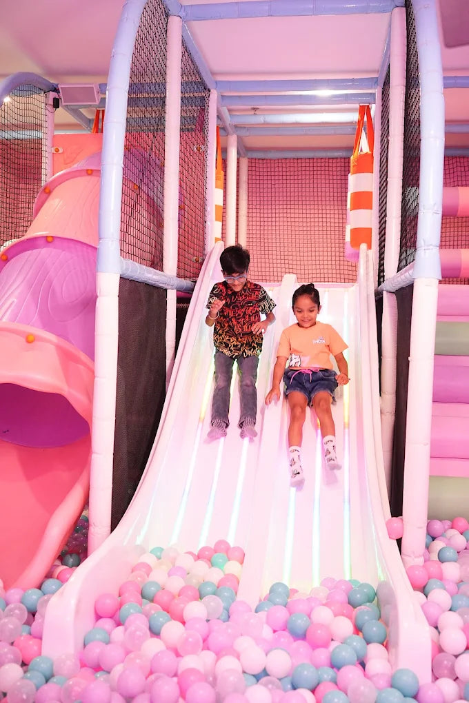 Two children slide down a colorful playground slide into a ball pit.