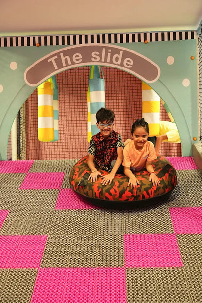 Two children sit in an inflatable tube at the bottom of a slide in a colorful indoor play area.