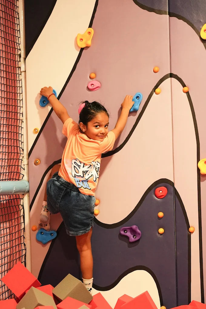 A young girl climbs a colorful indoor climbing wall, smiling at the camera. She is surrounded by soft foam blocks.