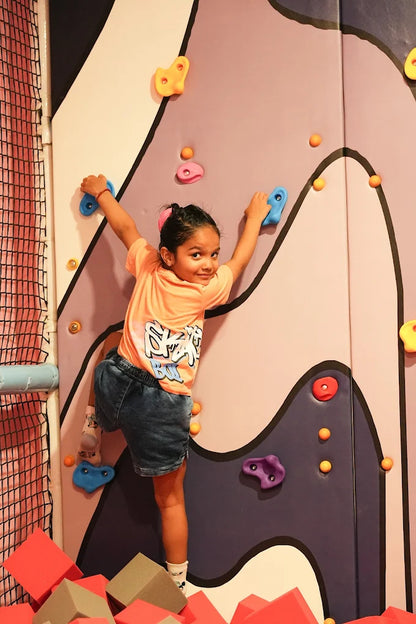 A young girl climbs a colorful indoor climbing wall, smiling at the camera. She is surrounded by soft foam blocks.