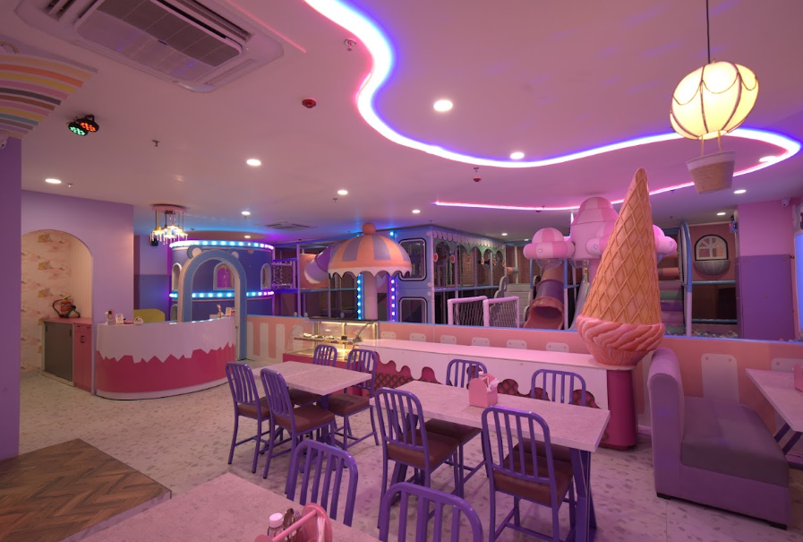 A brightly lit indoor playground with tables and chairs, designed for children. The space features colorful play structures and ice cream-themed decor.