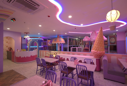 A brightly lit indoor playground with tables and chairs, designed for children. The space features colorful play structures and ice cream-themed decor.