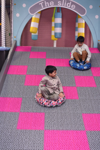 Two young boys slide down a colorful indoor playground slide on inflatable rings.