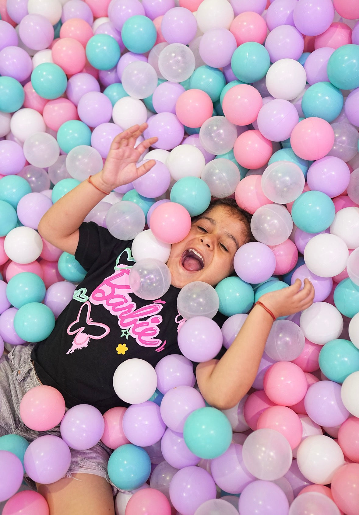 A young child joyfully lies in a ball pit filled with pastel-colored balls.