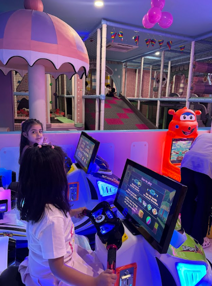 Two young girls play racing arcade games in a colorful indoor playground.