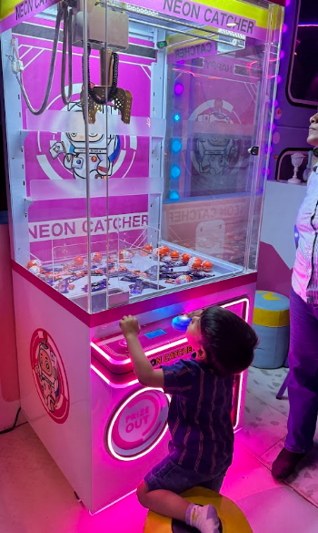A young child plays a neon pink claw machine game, reaching for prizes.