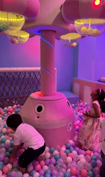 Children play in a ball pit with a large pink structure and colorful lights.