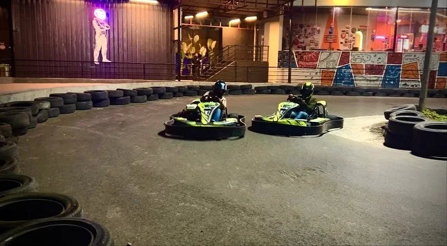 Two go-karts with drivers race on a track at night, with a neon astronaut figure visible in the background.