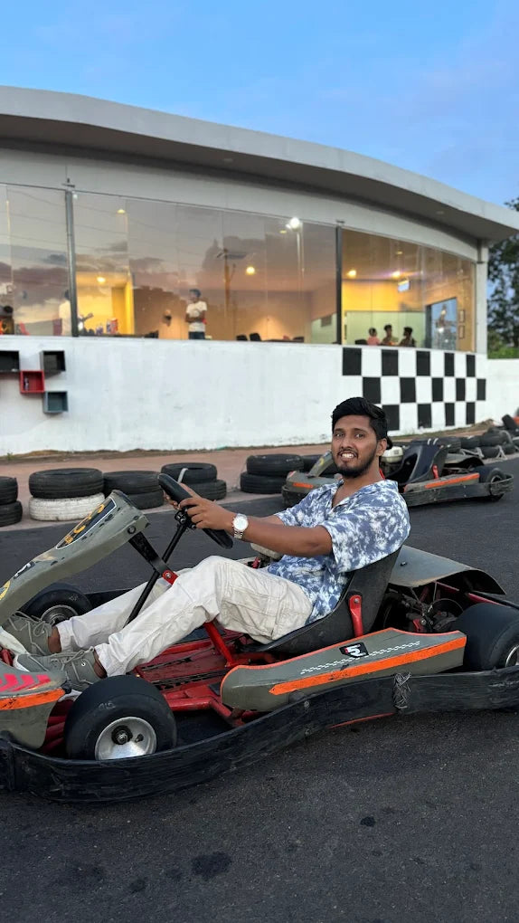 A young man smiles while sitting in a go-kart on a track, with a building in the background.