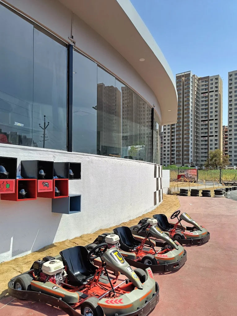 Go-karts lined up on a track in front of a building with large windows, with apartment buildings in the background.