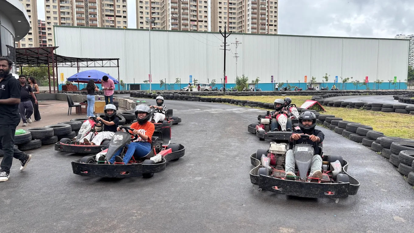 Young adults race go-karts on a track with tall buildings in the background.