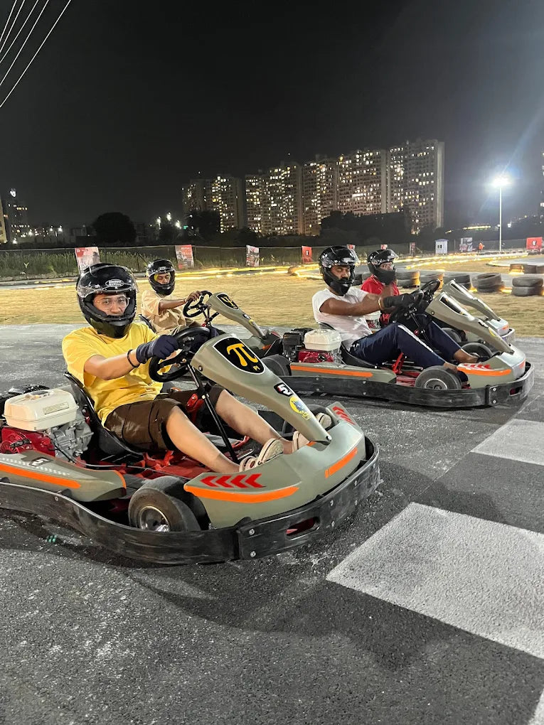 Adults race go-karts at night on a track with city lights in the background.