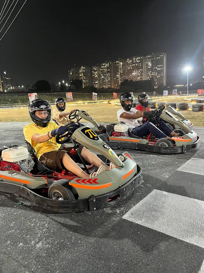 Adults race go-karts at night on a track with city lights in the background.