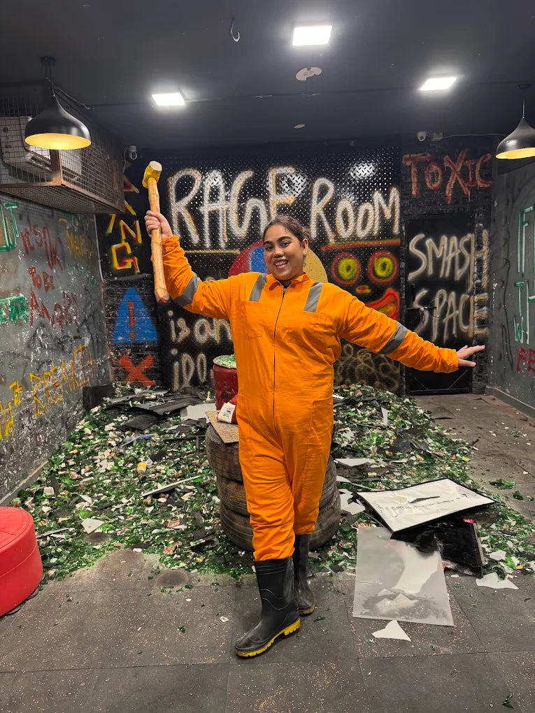 A young woman in an orange jumpsuit holds a sledgehammer in a rage room filled with broken glass.