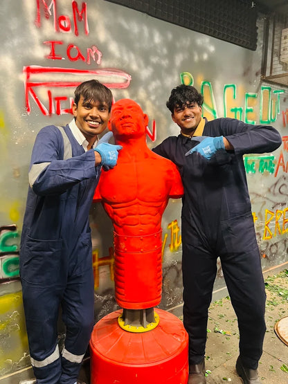 Two young men in blue jumpsuits pose with a red punching dummy in a graffiti-covered room.