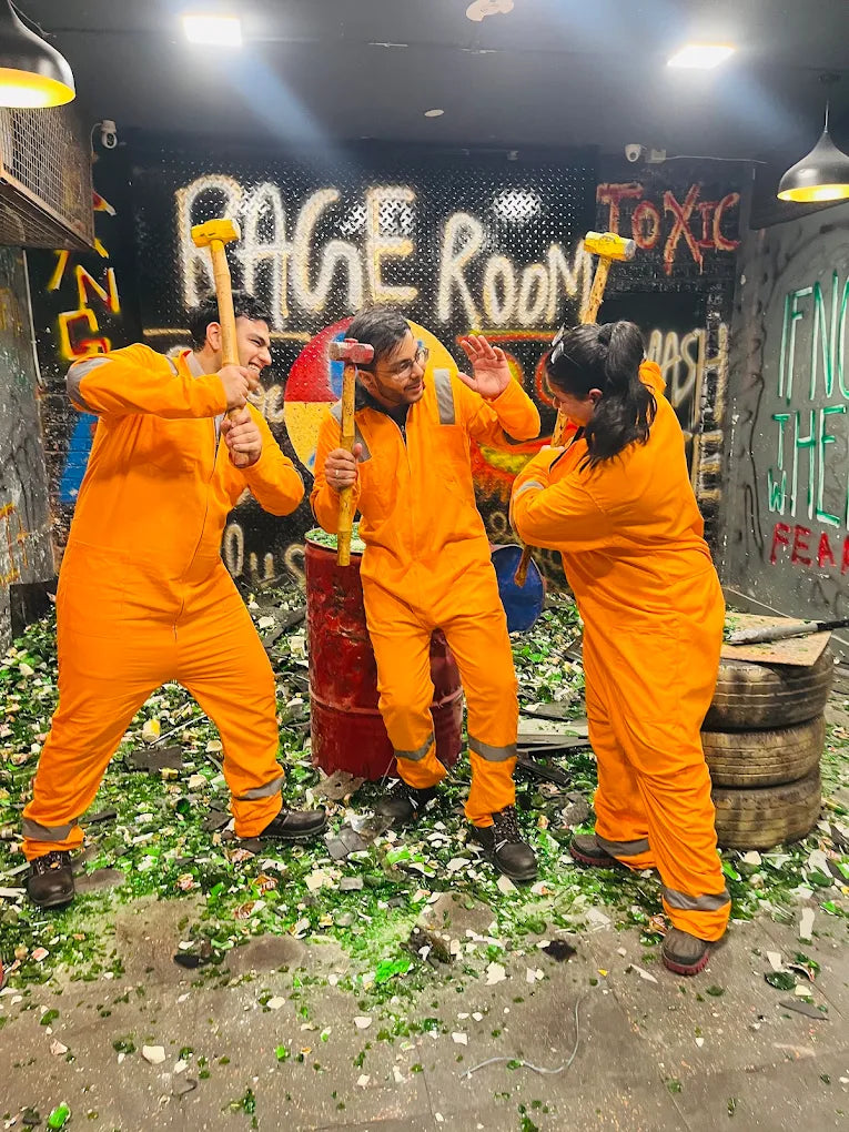 Three young adults in orange jumpsuits smash bottles with hammers in a rage room.