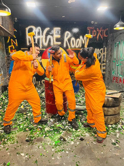 Three young adults in orange jumpsuits smash bottles with hammers in a rage room.