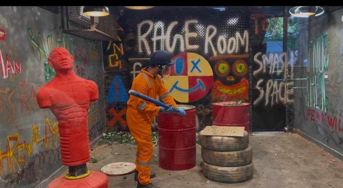 A person in an orange jumpsuit and helmet swings a bat in a graffiti-covered "Rage Room" filled with barrels and tires.
