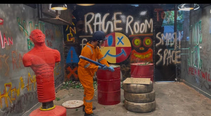 A person in an orange jumpsuit and helmet swings a bat in a graffiti-covered "Rage Room" filled with barrels and tires.