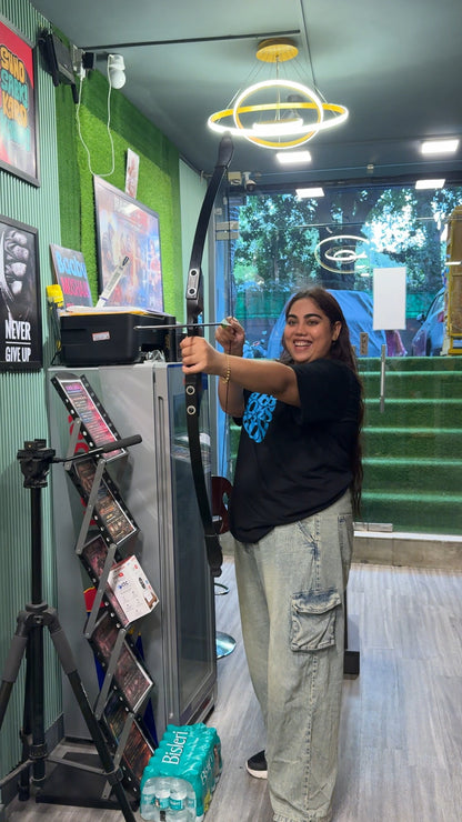 A young woman aims a bow and arrow indoors, smiling. She is standing in front of a refrigerator and a display rack.