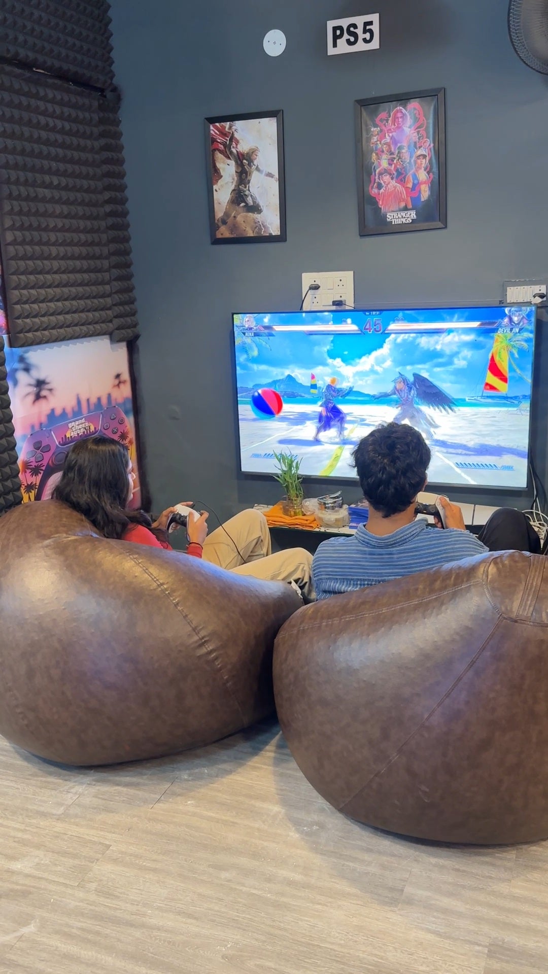 Two young adults play a video game on a large screen while sitting on beanbag chairs in a room with gaming posters.