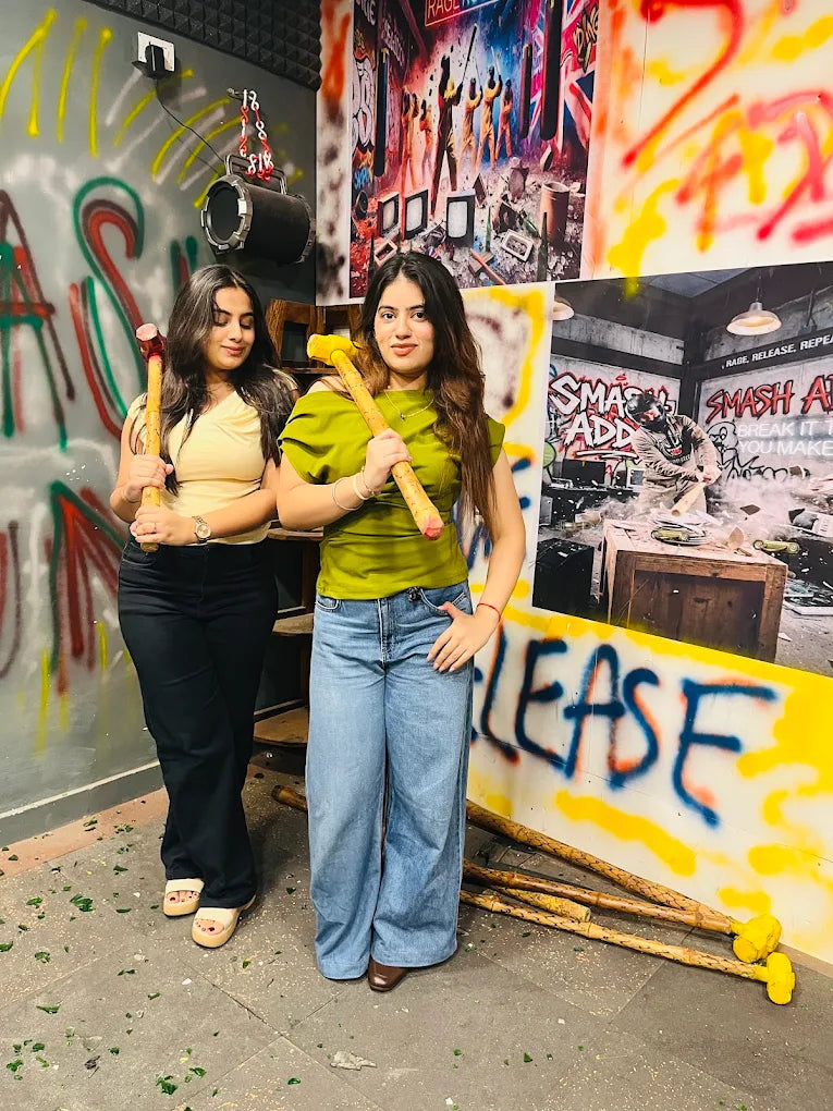Two young women hold sledgehammers in a room decorated with graffiti and broken glass.