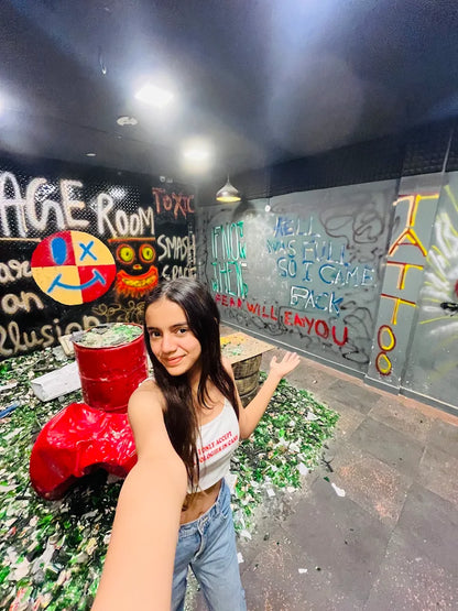 A young woman poses in a room filled with broken glass, surrounded by graffiti.