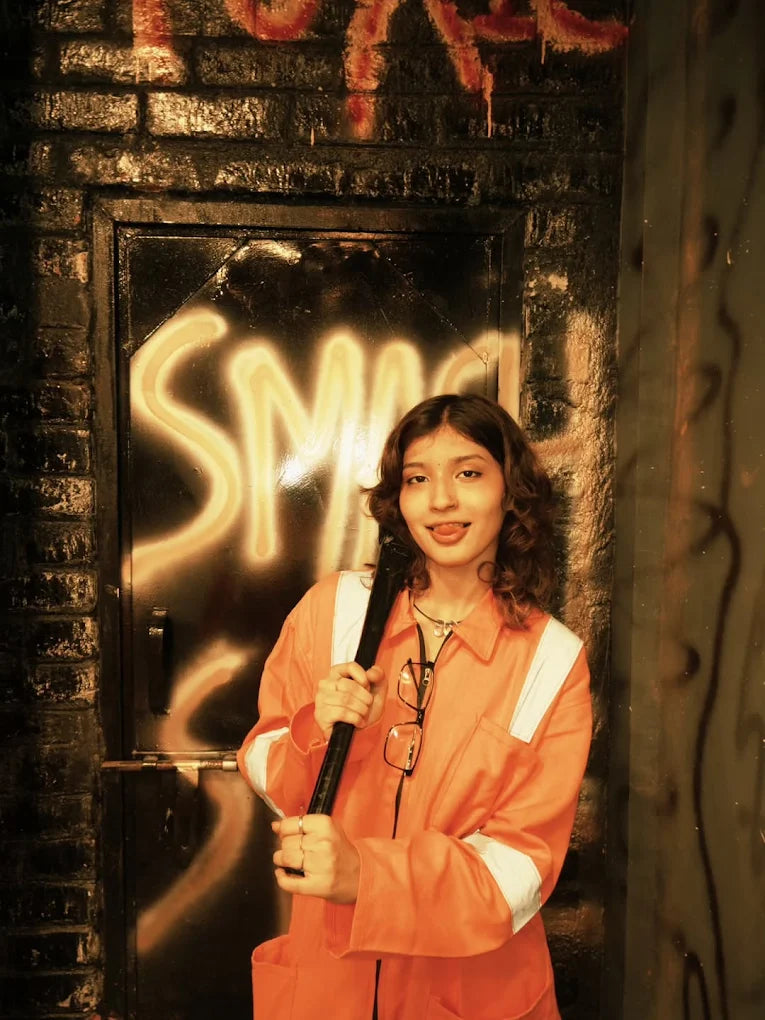 A young woman in an orange jumpsuit holds a bat in front of a graffiti-covered door.
