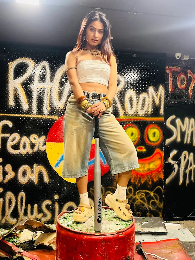 A young woman stands on a barrel holding a baseball bat in a graffiti-covered room.