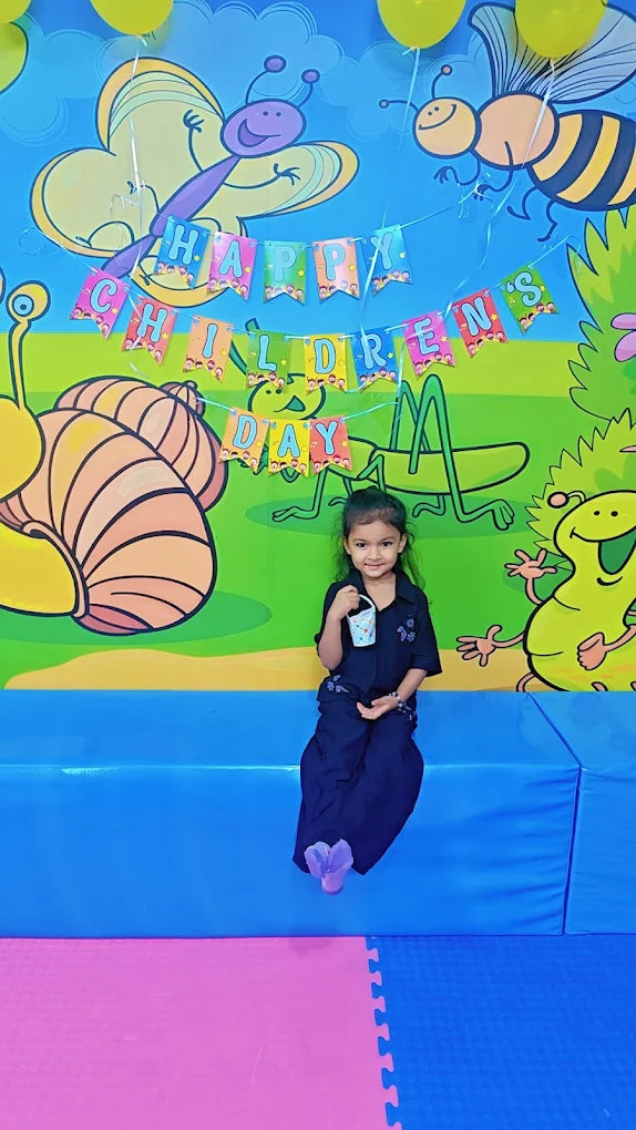 A young girl in a dark outfit sits in front of a colorful backdrop with "Happy Children's Day" banners.