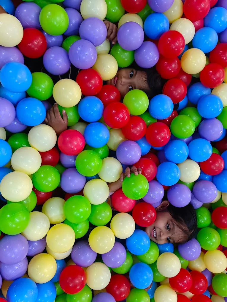 A young child peeks out from a ball pit filled with colorful plastic balls.