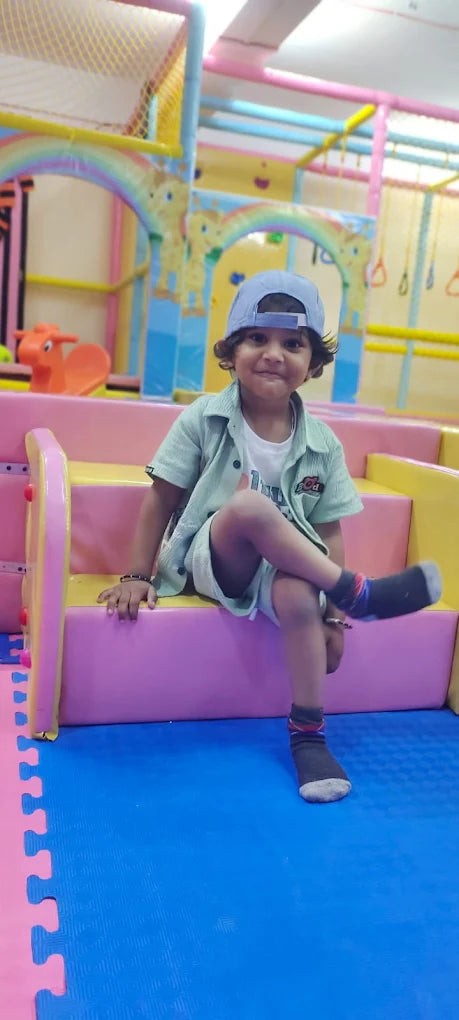 A young child sits on colorful steps in a play area, wearing a cap and a light green shirt.
