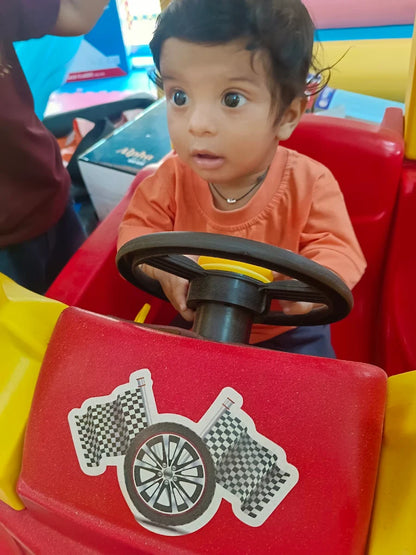 A baby sits in a toy car, holding the steering wheel. The car is red and yellow with racing flag stickers.