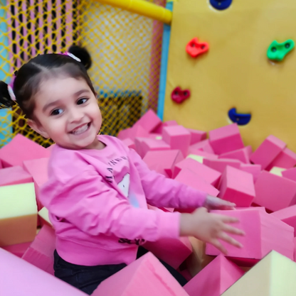 A young girl smiles while playing in a ball pit filled with pink and yellow foam blocks.