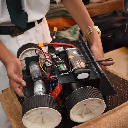 Student holding a small robot or electronic device with wheels on a wooden surface.
