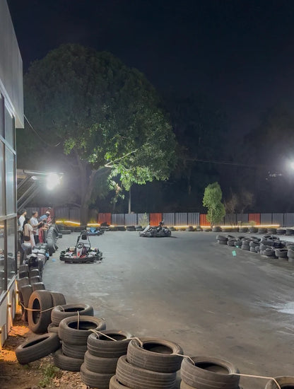 Adults race go-karts at a nighttime outdoor track lined with tires and trees.