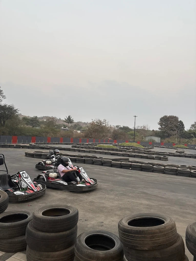 People race go-karts on a track lined with tires, with trees and a cloudy sky in the background.