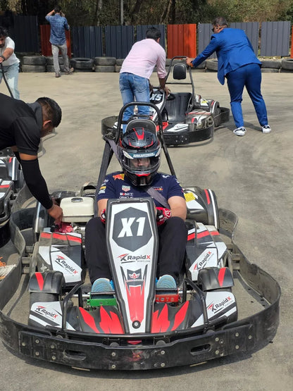 A young man in a racing helmet sits in a go-kart, ready to race. Other people are visible in the background at the outdoor track.