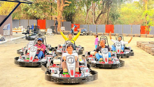 Adults in go-karts at an outdoor track, some with hands raised in excitement.