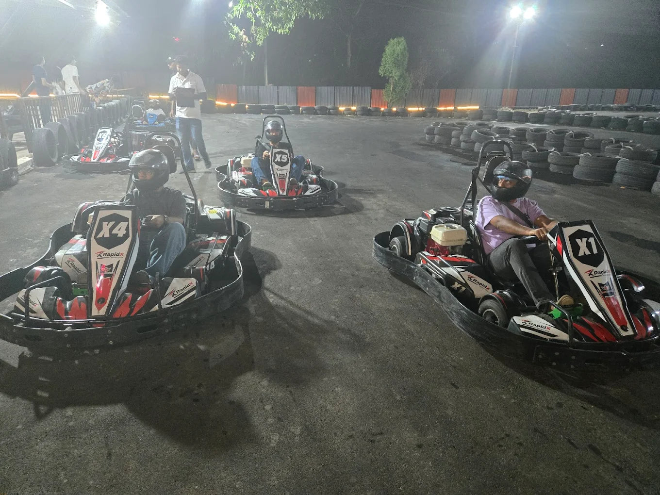 Adults race go-karts on a track at night.