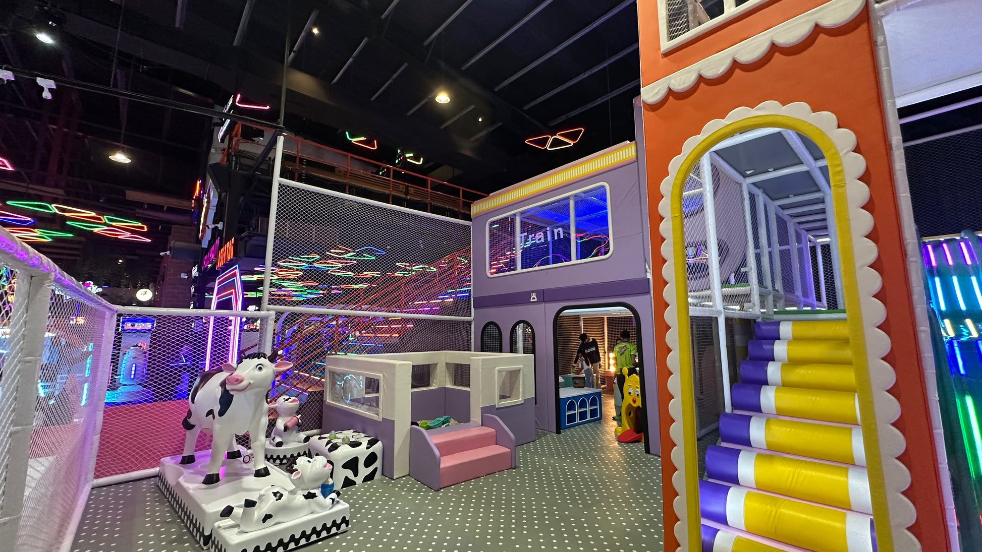 A colorful indoor playground with children playing, featuring a train-themed structure and cow decorations.