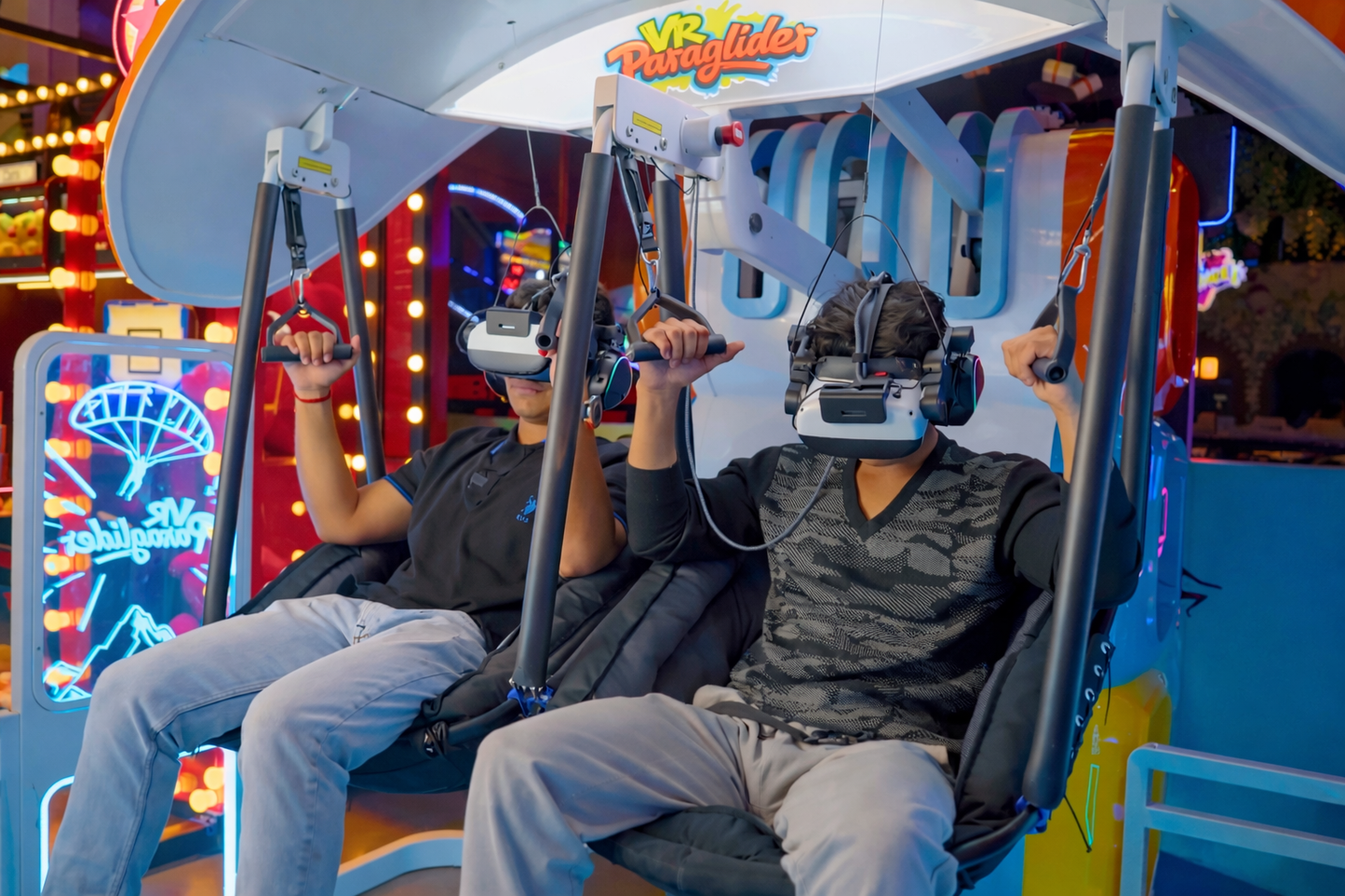 Two young people wearing VR headsets experience a virtual reality paraglider ride in an amusement park.