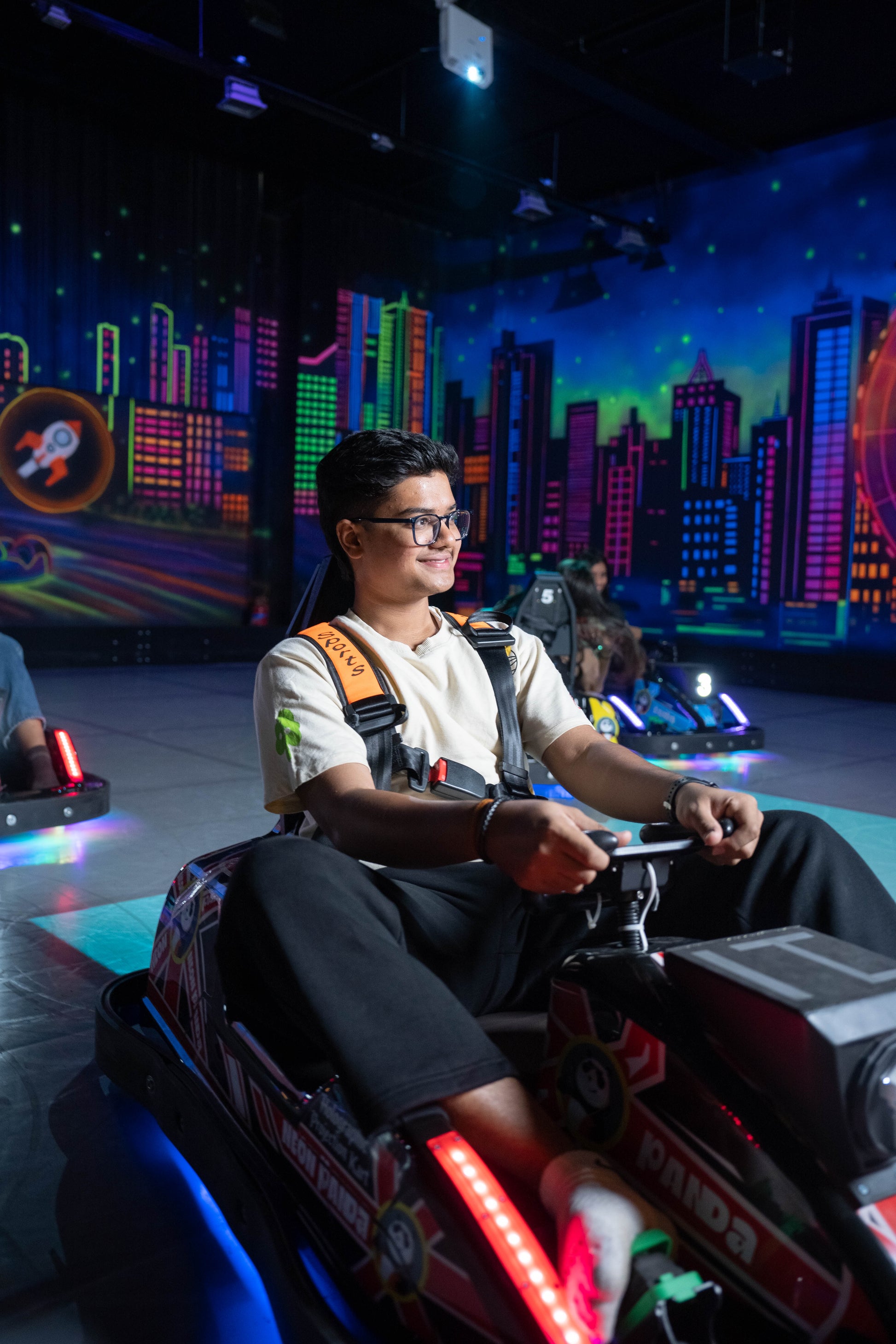 A young man smiles while driving a go-kart in an arcade with a neon city backdrop.