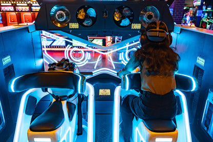 A person in VR gear sits in a futuristic arcade ride with bright blue lights. The ride features two seats and a large screen displaying neon graphics.